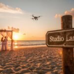 A Sri Lankan beach wedding at sunset with a couple under a floral arch, guests in sarees, and a drone above. The Studio Lanka logo is etched as a blueprint watermark on a signpost, in a pink-gold palette with lens flares and shallow depth of field.