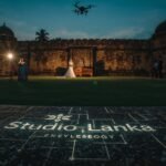 A Galle Fort scene at dusk with a film crew capturing a wedding video, a drone hovering, and a couple posing against colonial walls. The Studio Lanka logo is etched as a blueprint watermark on a stone bench, glowing with white technical linework. The blue-gold palette highlights fabrics and stone, with lens flares and a shallow depth of field.