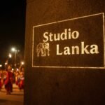 A Kandy Esala Perahera scene at night with a videographer filming vibrant dancers and elephants under torchlight. The Studio Lanka logo is etched as a blueprint watermark on a stone pillar, glowing with white technical linework. The red-gold palette highlights costumes, with fire glow and a shallow depth of field.